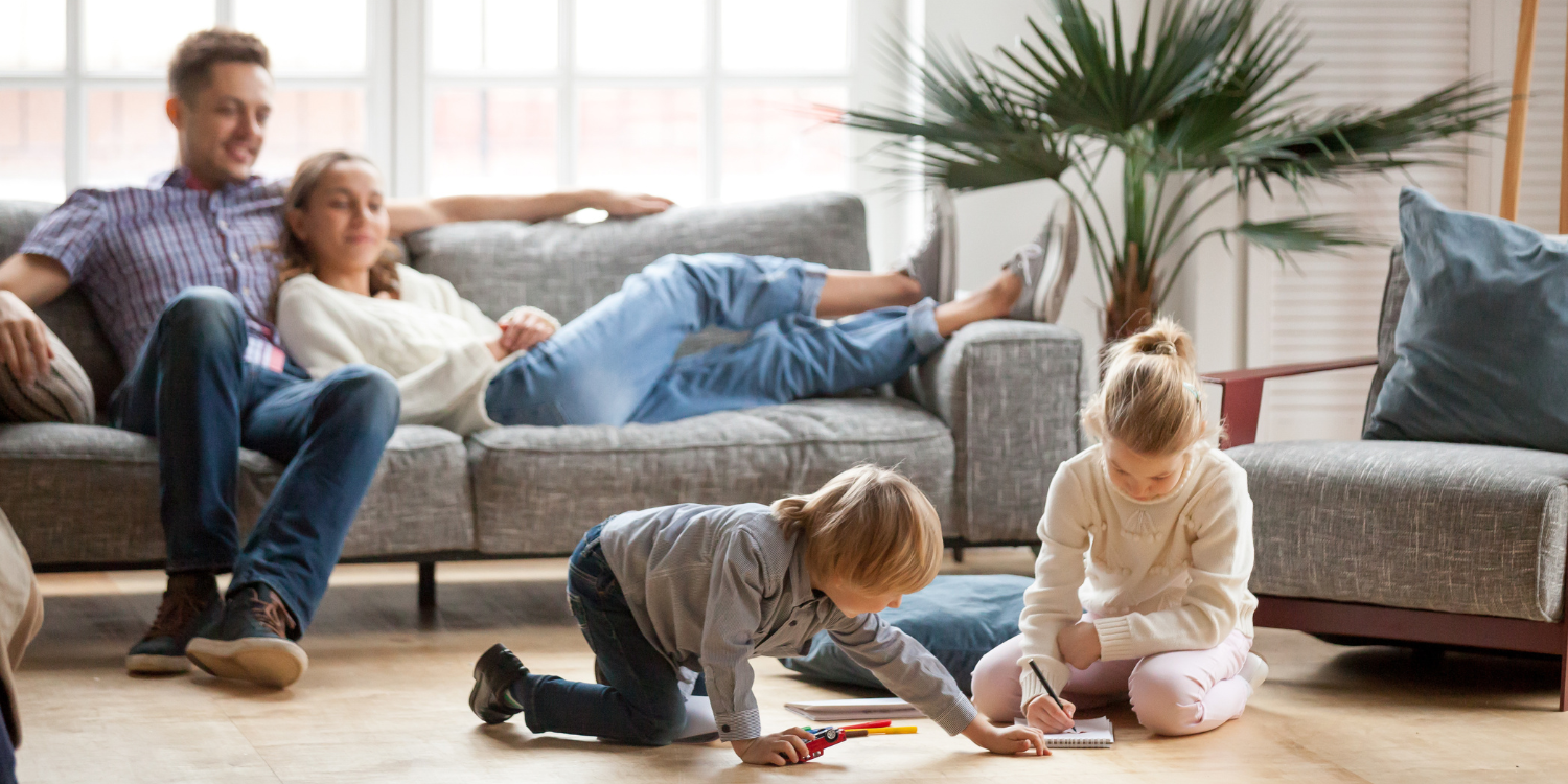 Family at home, parents on coach watching kids play with toys - Now’s the Perfect Time to Book Your Furnace Tune-Up. Here’s Why!