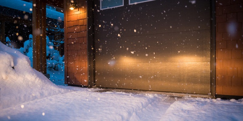 Car headlights shining into closed garage door, snow falling and snow on the ground